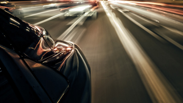 Automóvil en movimiento captado de noche, con efecto de desenfoque y estelas de luces de otros vehículos y farolas que sugieren velocidad en una carretera urbana.