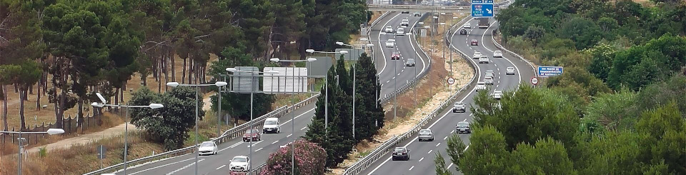 Vista aérea de una carretera de acceso a una ciudad con dos carriles por sentido, tráfico denso de vehículos y árboles alineados a los lados Vista aérea de una carretera de acceso a una ciudad con dos carriles por sentido, tráfico denso de vehículos y árboles alineados a los lados