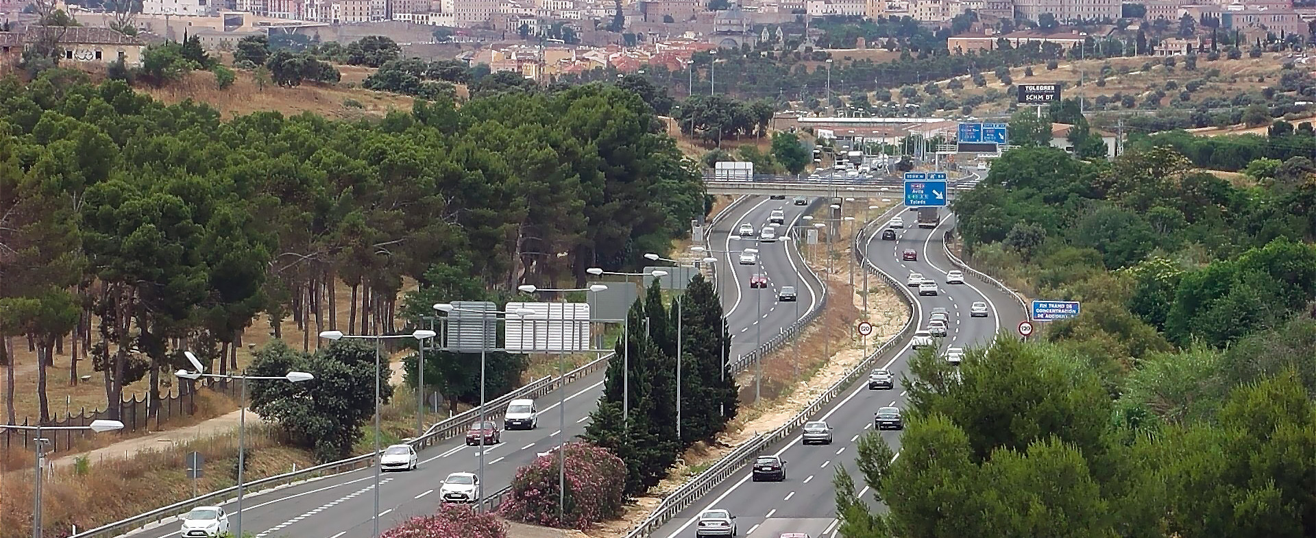 Vista aérea de una carretera de acceso a una ciudad con dos carriles por sentido, tráfico denso de vehículos y árboles alineados a los lados