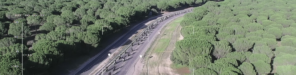 Vista aérea de una carretera rodeada de un extenso bosque de pinos, con una larga fila de motocicletas avanzando en formación. En la imagen se muestran coordenadas y fecha en la esquina inferior Vista aérea de una carretera rodeada de un extenso bosque de pinos, con una larga fila de motocicletas avanzando en formación. En la imagen se muestran coordenadas y fecha en la esquina inferior