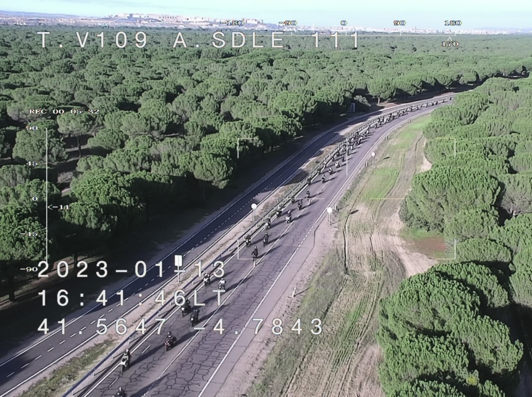 Vista aérea de una carretera rodeada de un extenso bosque de pinos, con una larga fila de motocicletas avanzando en formación. En la imagen se muestran coordenadas y fecha en la esquina inferior