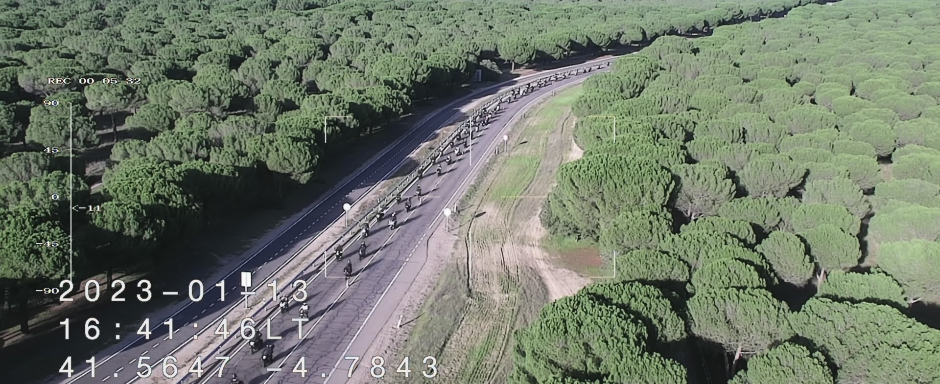 Vista aérea de una carretera rodeada de un extenso bosque de pinos, con una larga fila de motocicletas avanzando en formación. En la imagen se muestran coordenadas y fecha en la esquina inferior