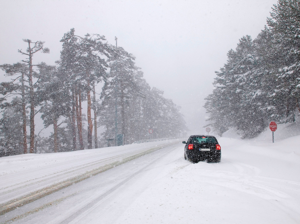 Coche negro detenido en medio de una nevada, con las luces de freno encendidas, en una carretera nevada rodeada de árboles muy altos al fondo