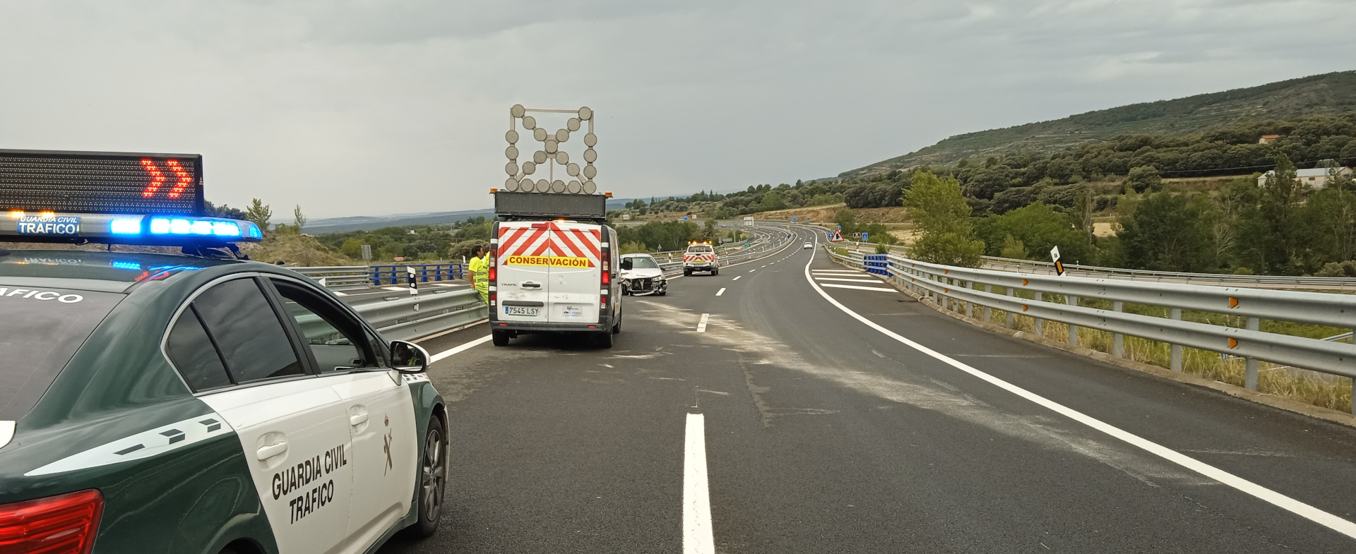 Carretera con curvas. A la izquierda, un coche de la Guardia Civil de Tráfico detenido. Al fondo, una furgoneta de Conservación de Carreteras está situada frente a un coche accidentado