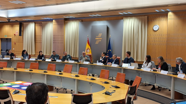 Una reunión de trabajo en una sala de conferencias semicircular con representantes institucionales y de seguridad sentados a una gran mesa. Al fondo, destacan las banderas de España y la Unión Europea junto a un logotipo institucional. Los asistentes tienen placas de identificación y micrófonos frente a ellos en un entorno profesional y formal.
