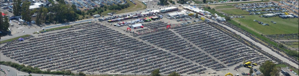 Imagen aérea de la explanada de una carretera con un parking donde se ven motocicletas aparcadas ordenadamente y unas carpas de eventos. También hay otros vehículos aparcados y vegetación.