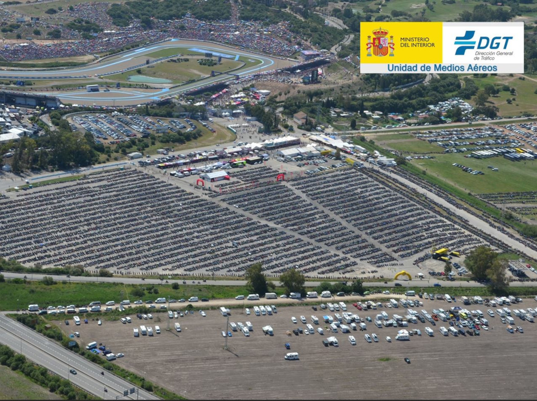 Imagen aérea de la explanada de una carretera con un parking donde se ven motocicletas aparcadas ordenadamente y unas carpas de eventos. También hay otros vehículos aparcados y vegetación. 