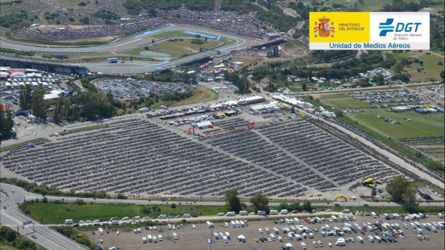 Imagen aérea de la explanada de una carretera con un parking donde se ven motocicletas aparcadas ordenadamente y unas carpas de eventos. También hay otros vehículos aparcados y vegetación.