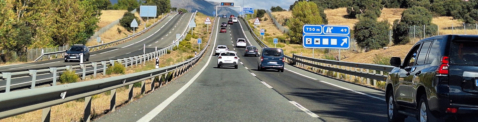 Vista frontal de una autovía de dos carriles por sentido con tráfico moderado. A los lados hay árboles y al fondo se ven montañas bajo un cielo azul con nubes.