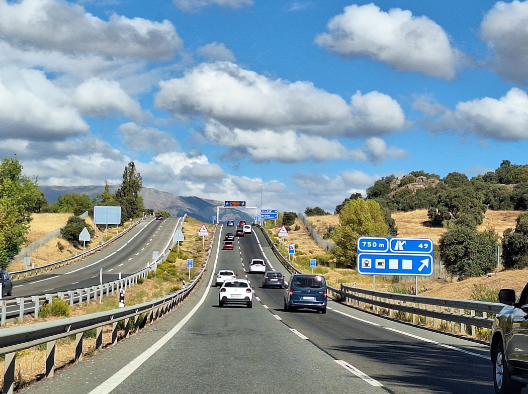 Vista frontal de una autovía de dos carriles por sentido con tráfico moderado. A los lados hay árboles y al fondo se ven montañas bajo un cielo azul con nubes.