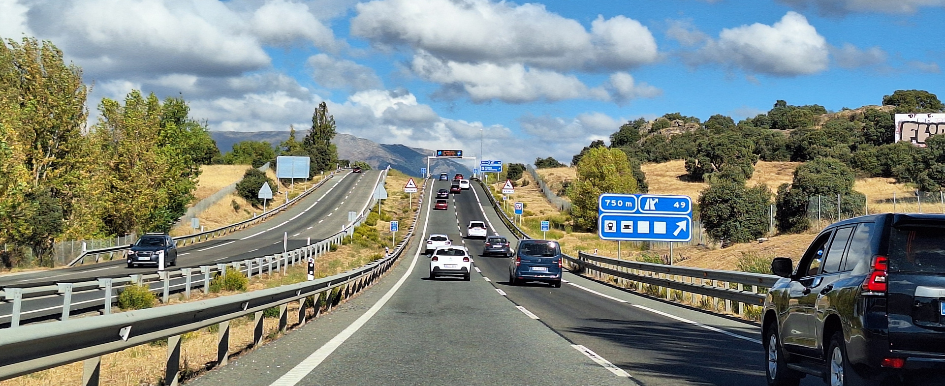 Vista frontal de una autovía de dos carriles por sentido con tráfico moderado. A los lados hay árboles y al fondo se ven montañas bajo un cielo azul con nubes.