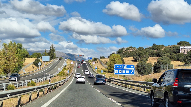 Vista frontal de una autovía de dos carriles por sentido con tráfico moderado. A los lados hay árboles y al fondo se ven montañas bajo un cielo azul con nubes.