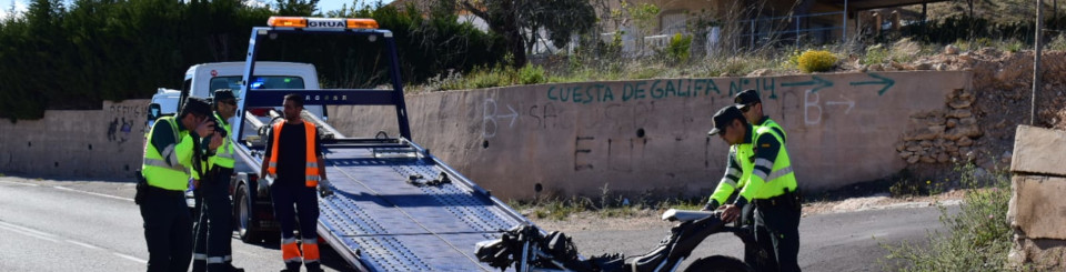 Accidente de motocicleta en una carretera. La moto está con daños visibles y sin la rueda delantera y la sujetan dos Guardias Civiles mientras al lado hay otros dos guardias, uno de ellos con una cámara en la mano haciendo fotografías a la moto. Al fondo hay una grúa con la rampa desplegada y un operario a su lado. 
