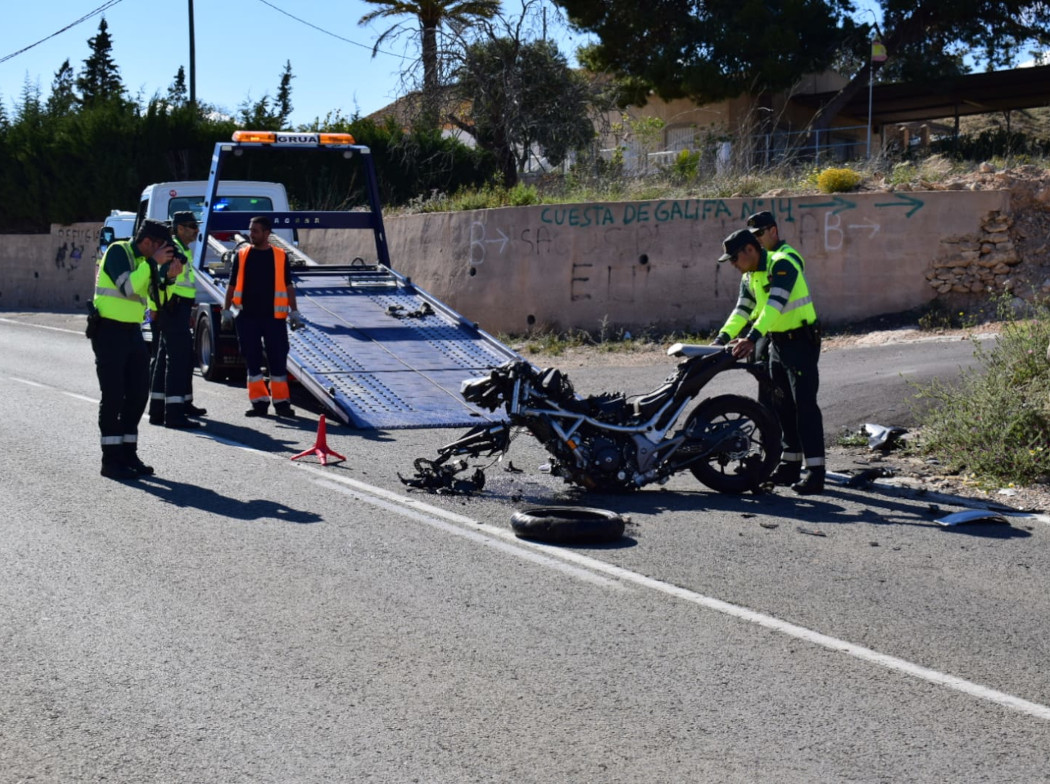 Accidente de motocicleta en una carretera. La moto está con daños visibles y sin la rueda delantera y la sujetan dos Guardias Civiles mientras al lado hay otros dos guardias, uno de ellos con una cámara en la mano haciendo fotografías a la moto. Al fondo hay una grúa con la rampa desplegada y un operario a su lado.