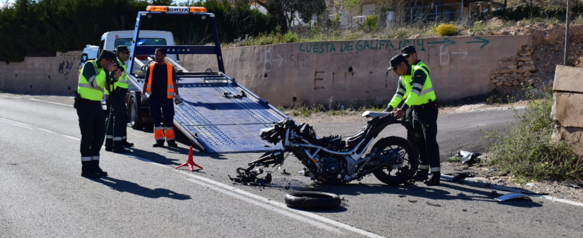 Accidente de motocicleta en una carretera. La moto está con daños visibles y sin la rueda delantera y la sujetan dos Guardias Civiles mientras al lado hay otros dos guardias. Al fondo hay una grúa con la rampa desplegada y un operario a su lado.