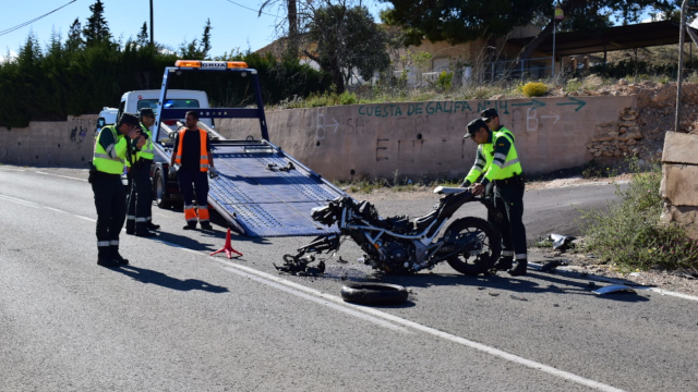 Accidente de motocicleta en una carretera. La moto está con daños visibles y sin la rueda delantera y la sujetan dos Guardias Civiles mientras al lado hay otros dos guardias. Al fondo hay una grúa con la rampa desplegada y un operario a su lado. 