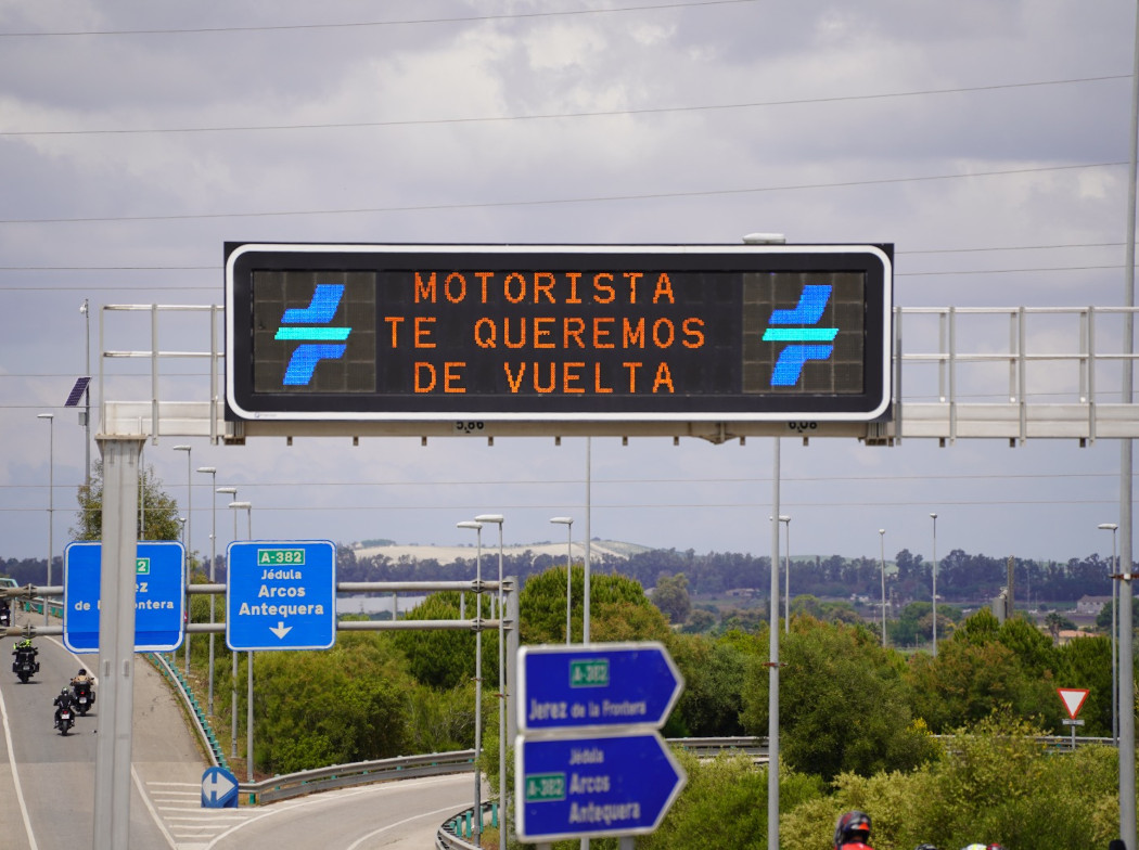 Fotografía de un cartel luminoso de carretera en el que se puede leer Motorista te queremos de vuelta. Justamente de bajo se ve a unos motoristas circulando por una autovía con carteles.