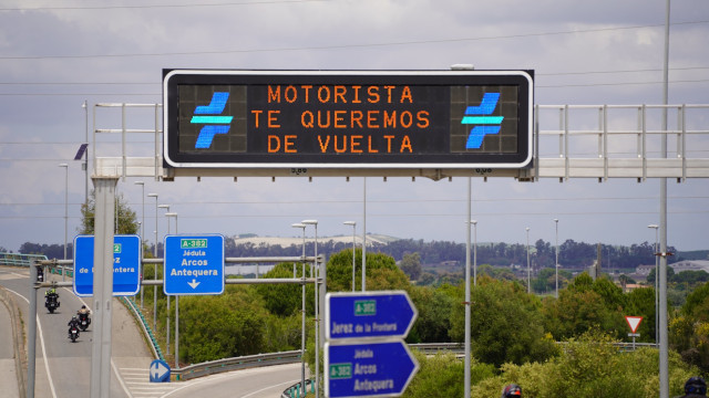 Fotografía de un cartel luminoso de carretera en el que se puede leer Motorista te queremos de vuelta. Justamente de bajo se ve a unos motoristas circulando por una autovía con carteles.
