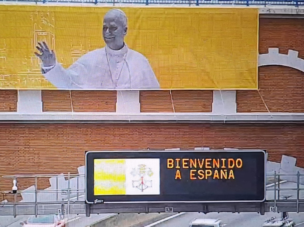 Cartel grande con la imagen del Papa León XIV desplegado en un puente de ladrillos. Debajo un panel de carretera con el texto "Bienvenido a España" y la bandera del Vaticano.