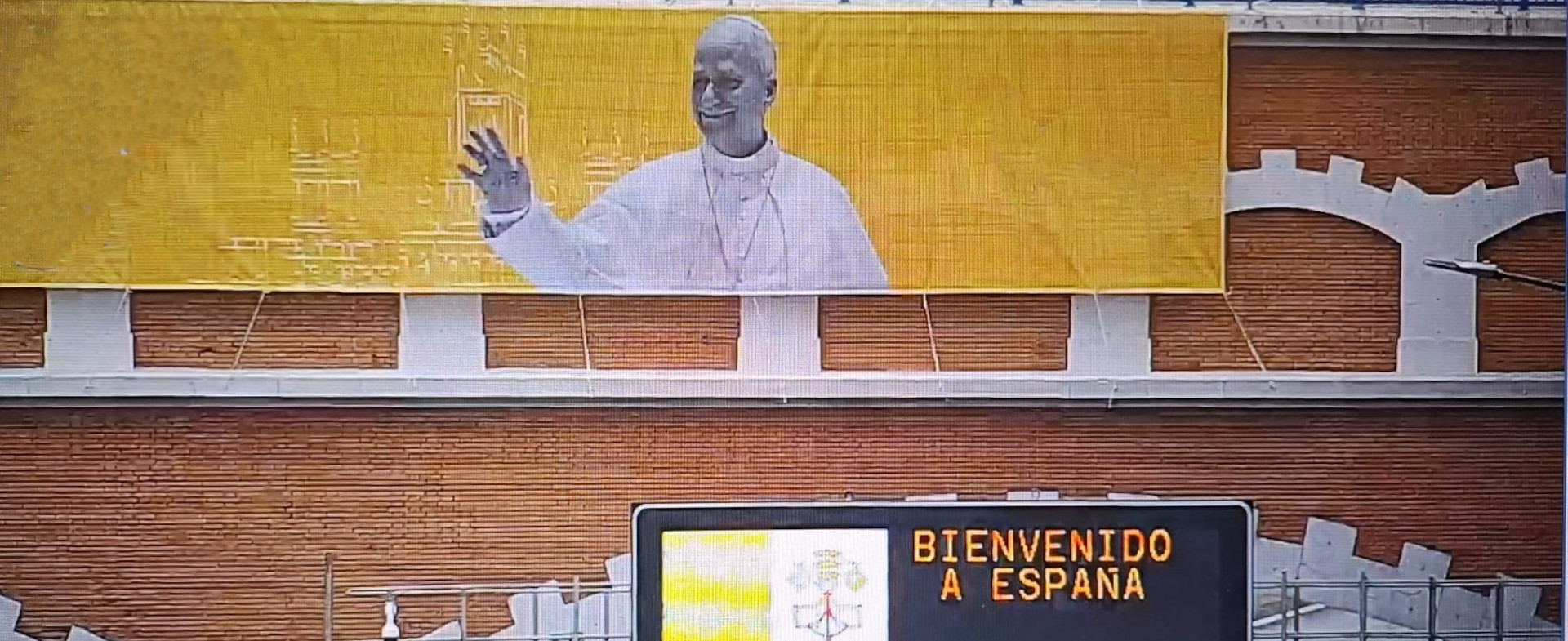 Cartel grande con la imagen del Papa León XIV desplegado en un puente de ladrillos. Debajo un panel de carretera con el texto "Bienvenido a España" y la bandera del Vaticano.