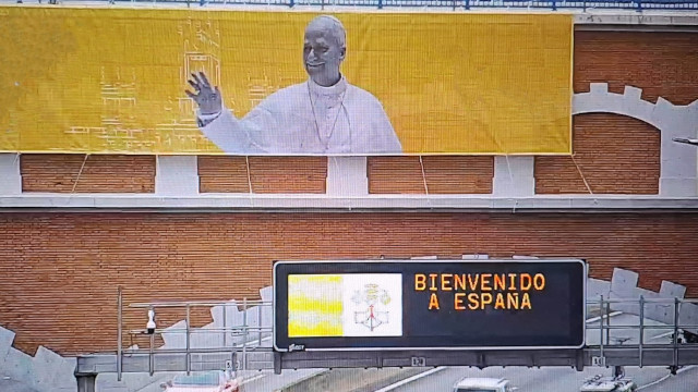 Cartel grande con la imagen del Papa León XIV desplegado en un puente de ladrillos. Debajo un panel de carretera con el texto 