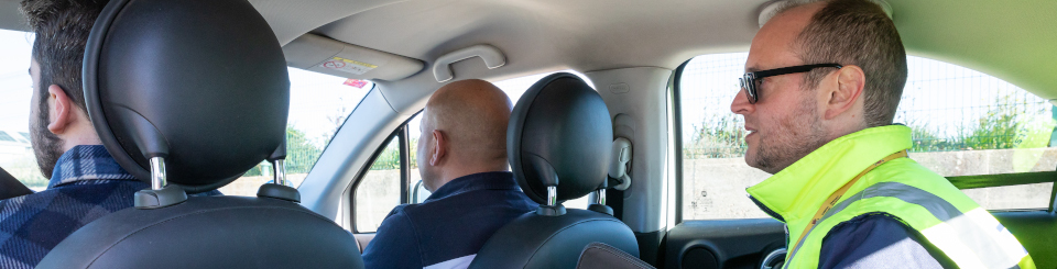 Hombre en el centro del asiento trasero de un coche, con carpeta y chaleco reflectante amarillo. Delante viajan dos hombres