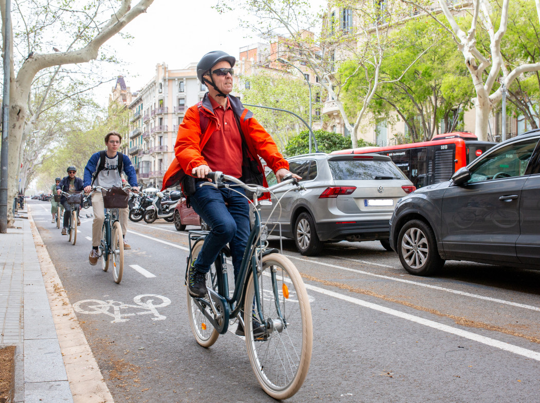 Tres ciclistas en hilera circulando por el carril bici en una calle de una ciudad. Al su lado, se encuentran coches aparcados.