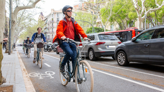 Tres ciclistas en hilera circulando por el carril bici en una calle de una ciudad. Al su lado, se encuentran coches aparcados.