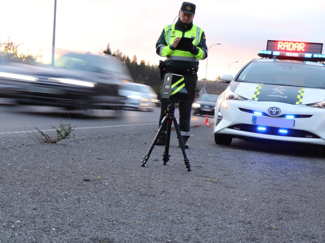 Agente de la Guardia Civil de Tráfico junto a un radar móvil en un trípode y su vehículo patrulla en la carretera.