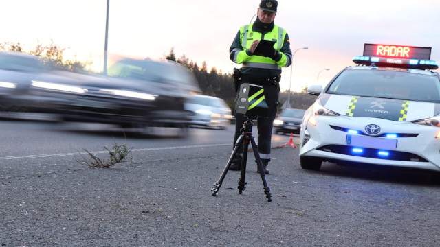 Agente de la Guardia Civil de Tráfico junto a un radar móvil en un trípode y su vehículo patrulla en la carretera.