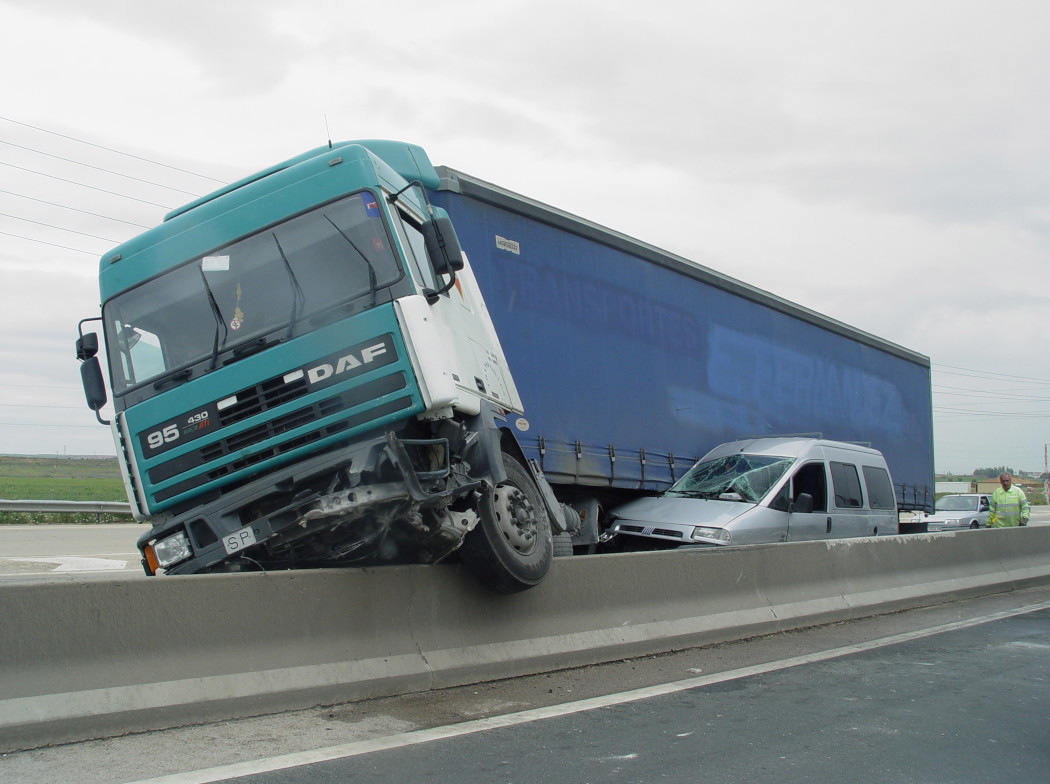 Camión accidentado sobre la barrera de una carretera, aplastando una furgoneta gris.