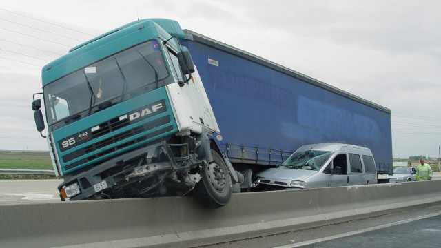 Camión accidentado sobre la barrera de una carretera, aplastando una furgoneta gris.