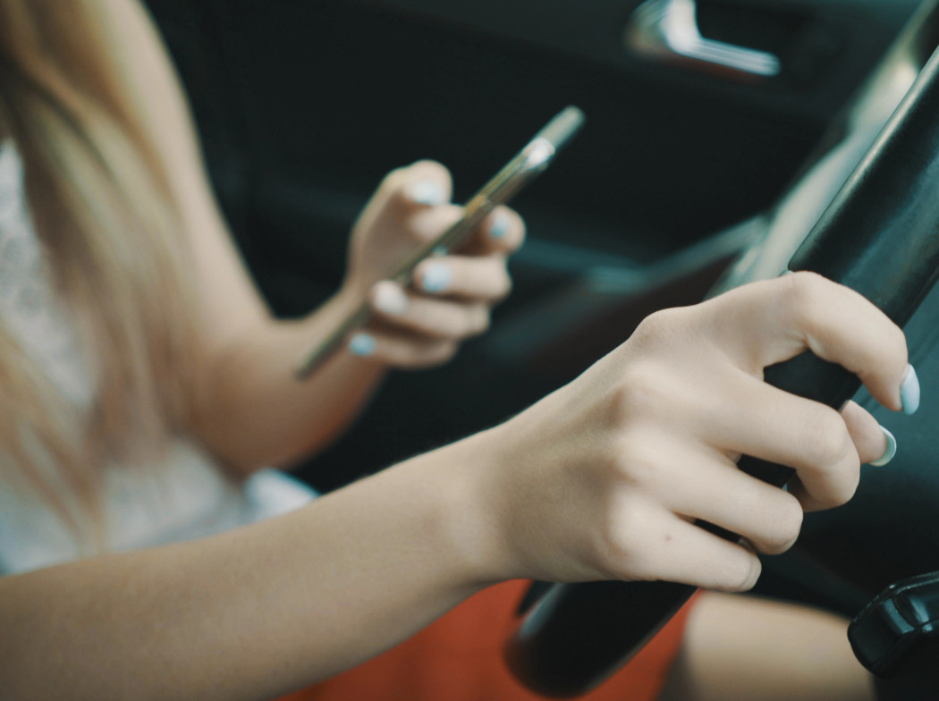 Mujer mirando el móvil en una mano y la otra puesta en el volante de un coche.