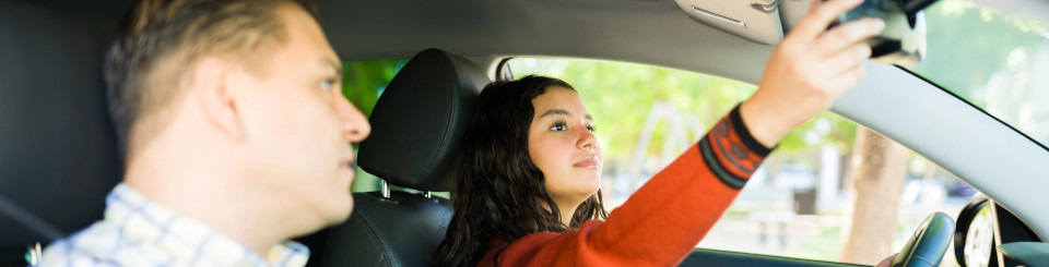 Una chica de 17 años dentro de un vehículo sentada el asiento del conductor, ajustando el espejo retrovisor. A su lado, un adulto señalando la ventana delantera.