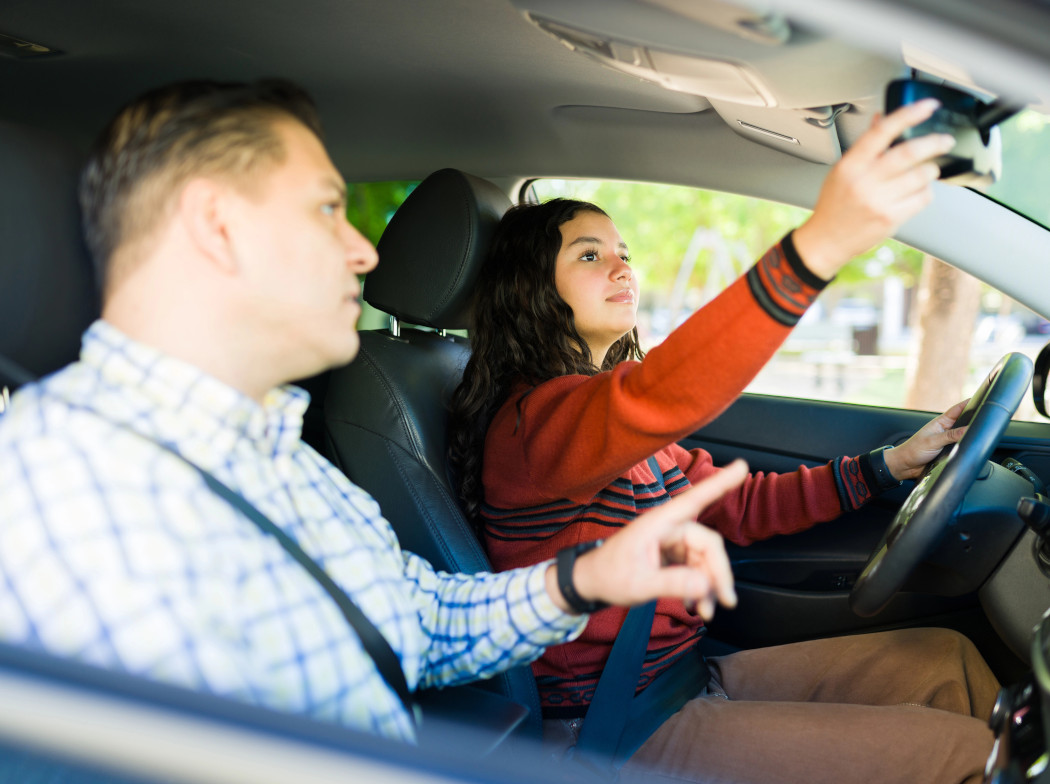 Una chica de 17 años dentro de un vehículo sentada el asiento del conductor, ajustando el espejo retrovisor. A su lado, un adulto señalando la ventana delantera.