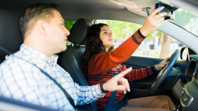 Una chica de 17 años dentro de un vehículo sentada el asiento del conductor, ajustando el espejo retrovisor. A su lado, un adulto señalando la ventana delantera.