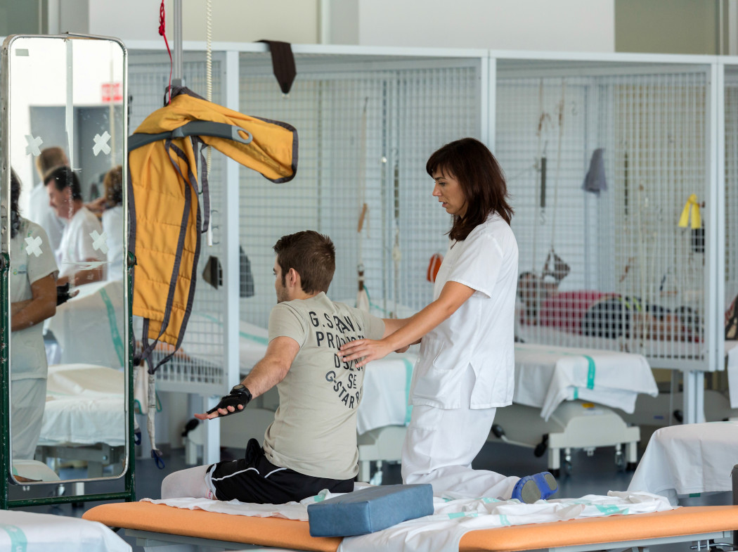 Hombre en rehabilitación en un centro con más pacientes haciendo estiramientos sentado en una cama mientras una enfermera le ayuda de pie a su lado.