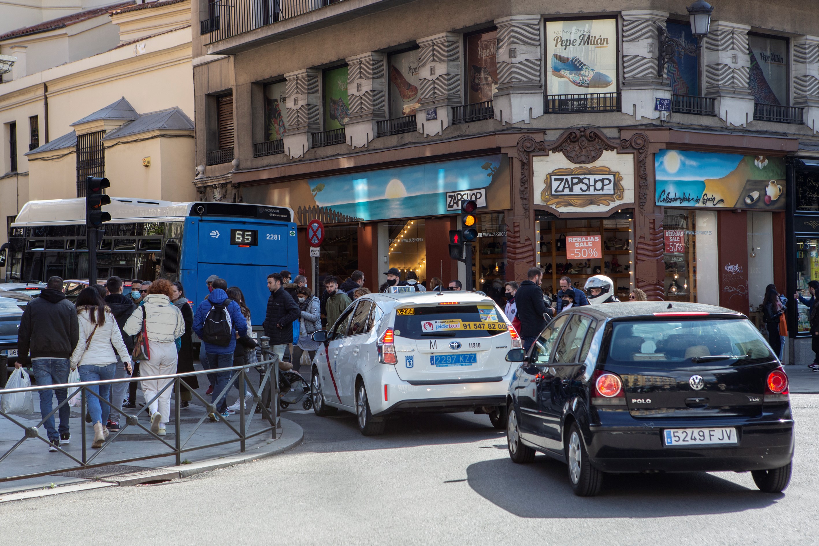 Imagen de calle de ciudad con transeúntes y cruzando un paso de peatones y varios vehículos parados dejando pasar.