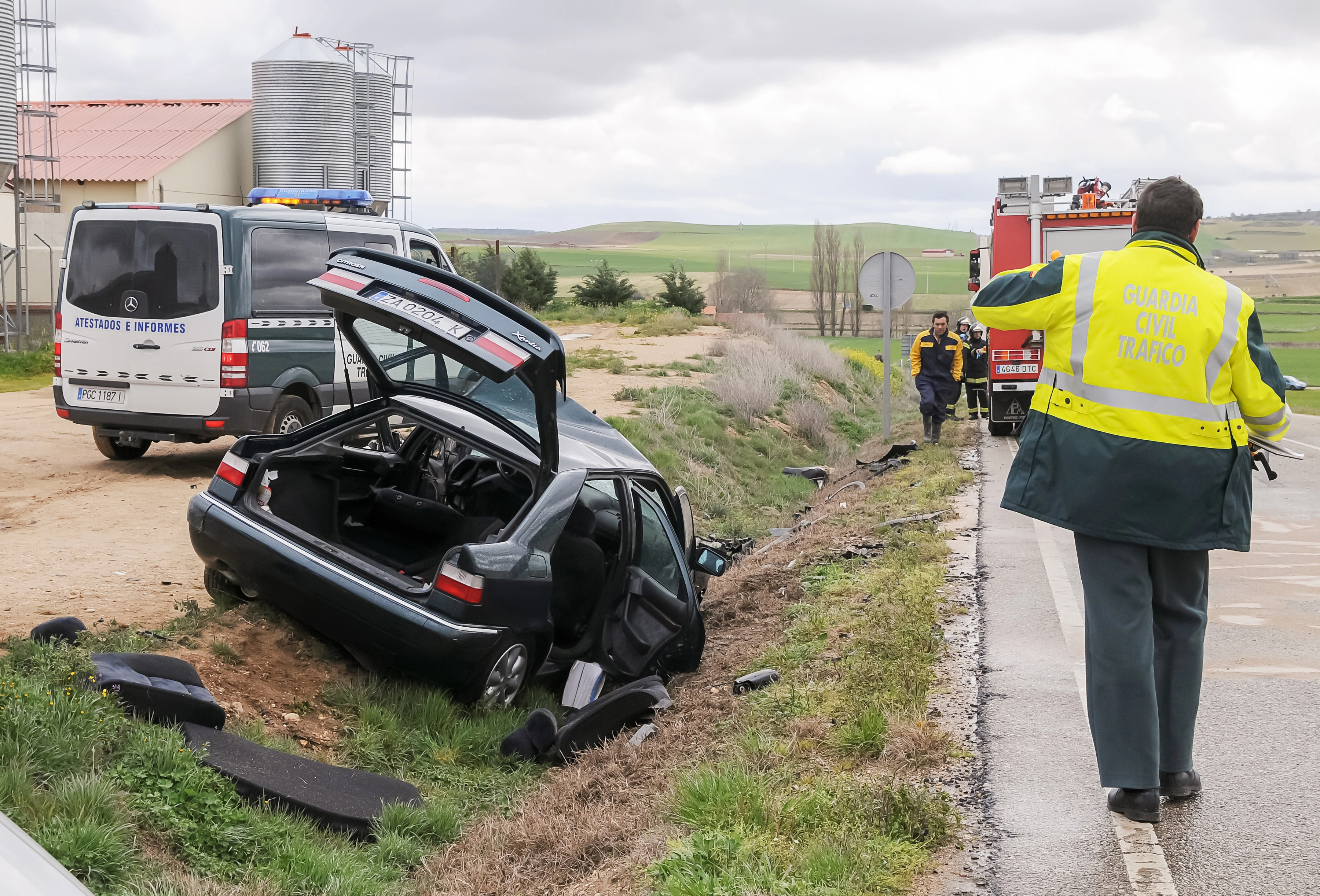 Coche accidentado en la cuneta de una carretera. A la derecha un hombre de la Guardia Civil andando por la carretera dirección a un coche de bomberos.