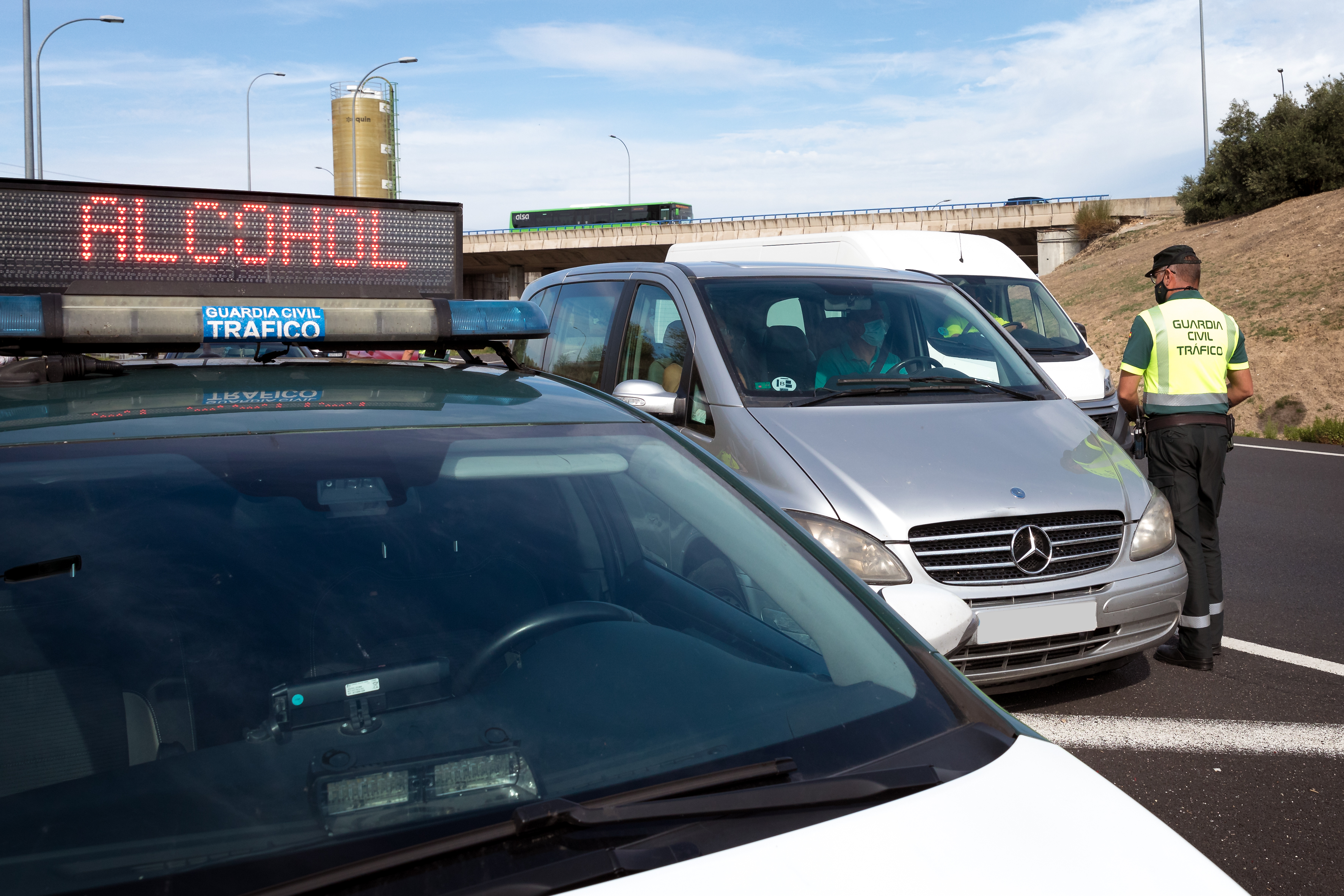 Coche de la Guardia Civil con un cartel lumínico en la parte superior del capó en el que pone "Alcohol". A su lado un Guardia Civil cerca de un coche.
