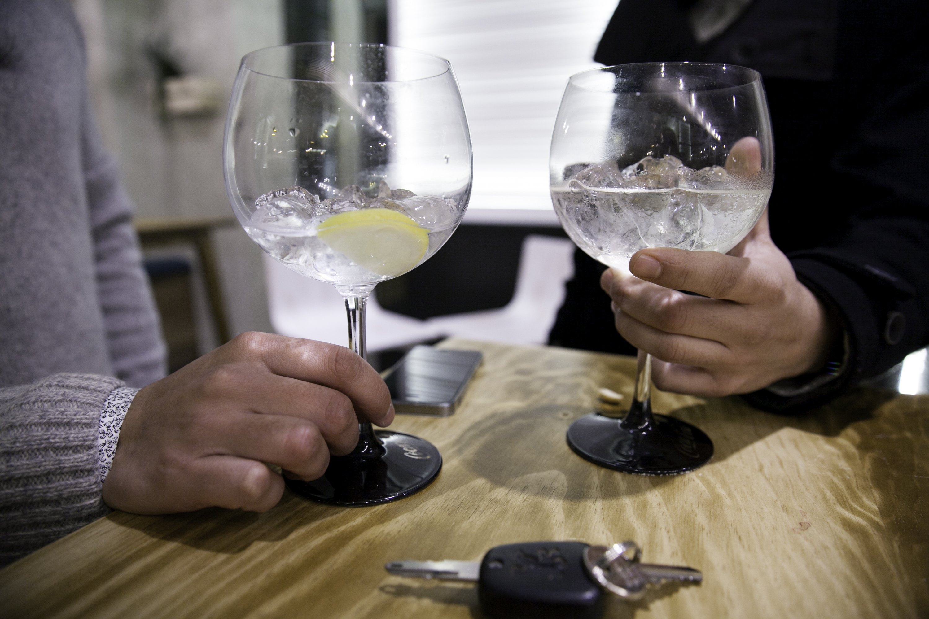 Dos manos masculinas sosteniendo dos copas de balón con hielos, limón y líquido transparente en su interior sobre una mesa. Delante de las copas hay unas llaves de un coche.
