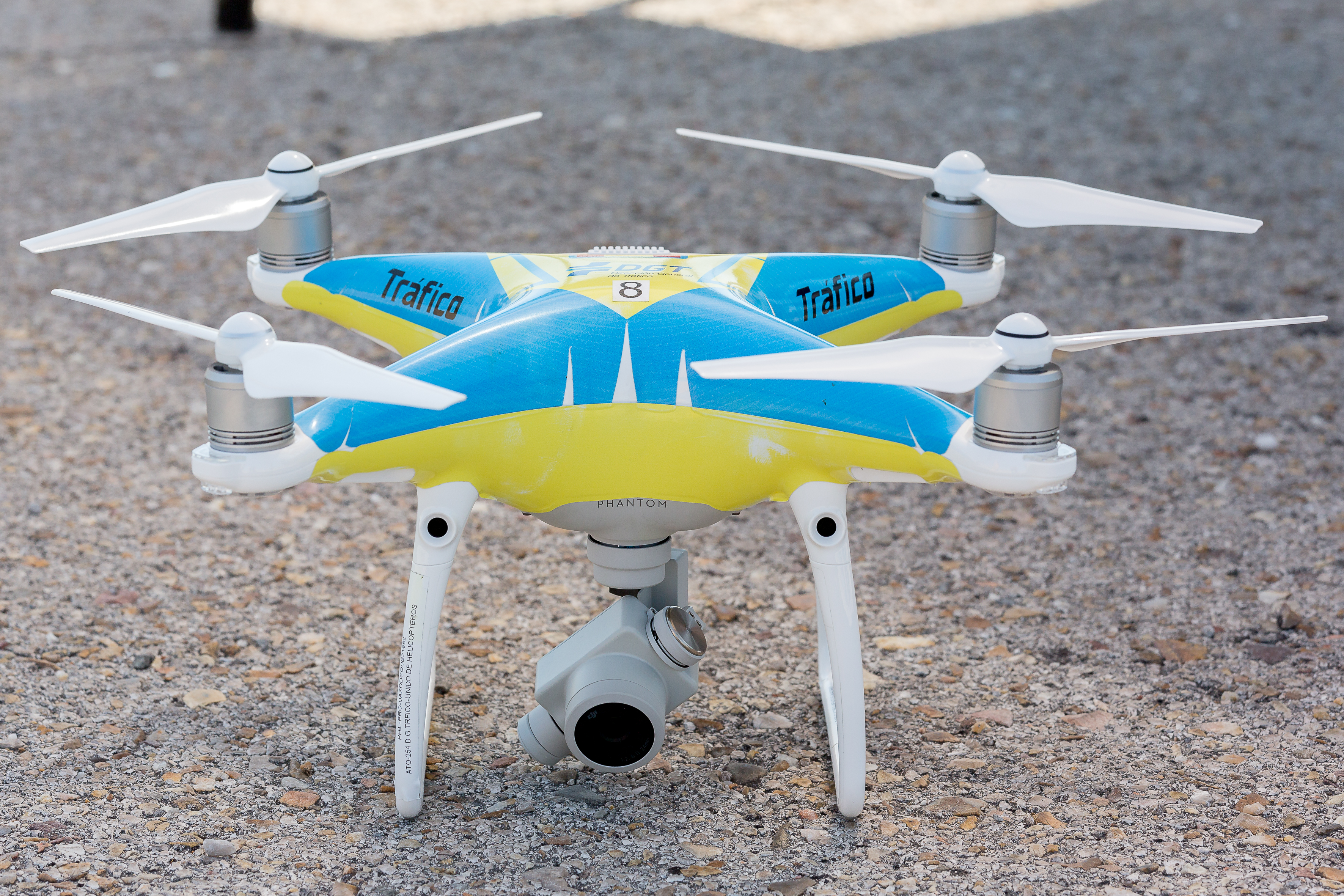 Un dron de cuatro hélices y cámara en la parte inferior con los colores amarillo y azul. Se encuentra parado en el suelo