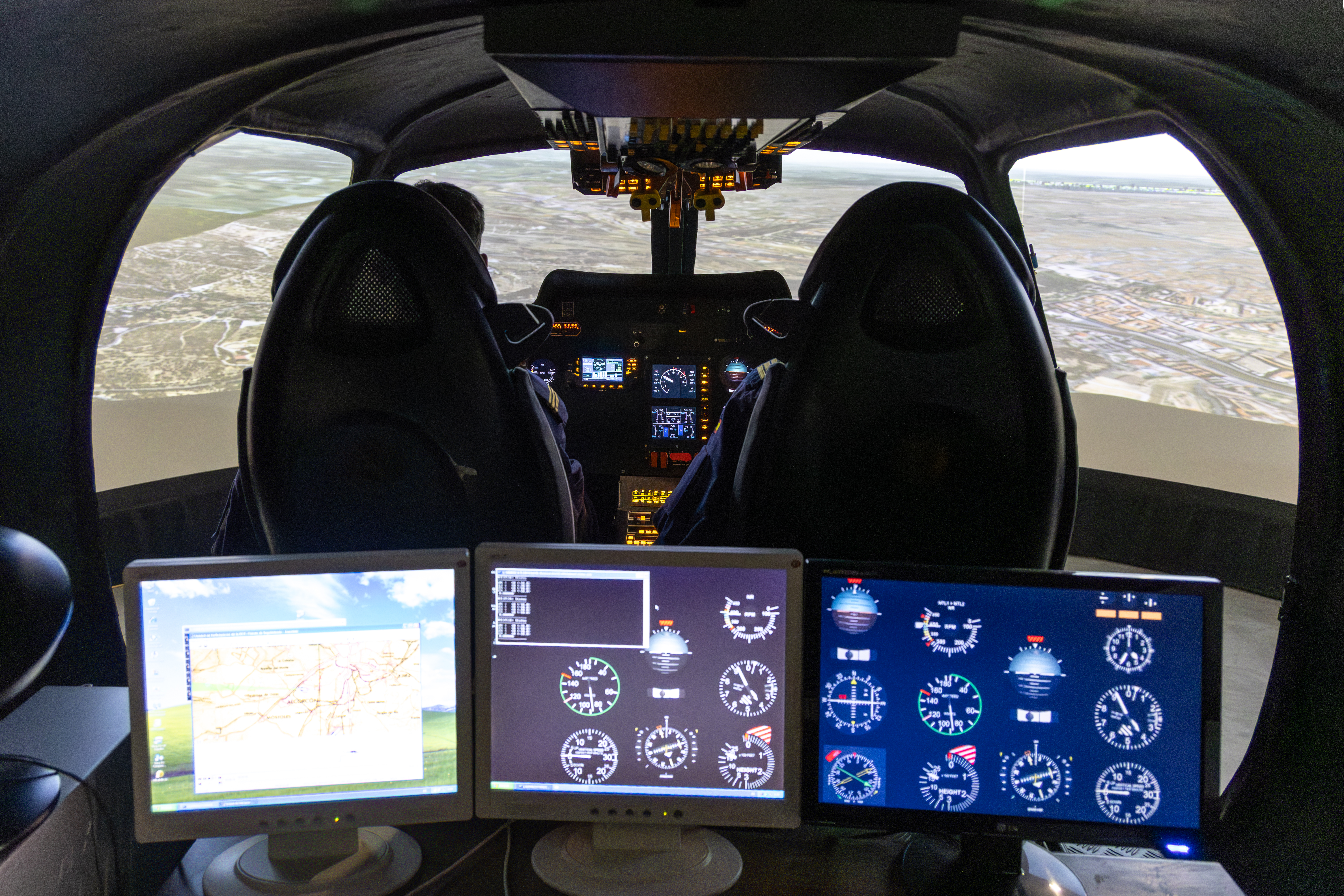 Simulador del interior de un helicóptero dónde hay tres pantallas encendidas y delante de ellas dos asientos junto con el tablero de control