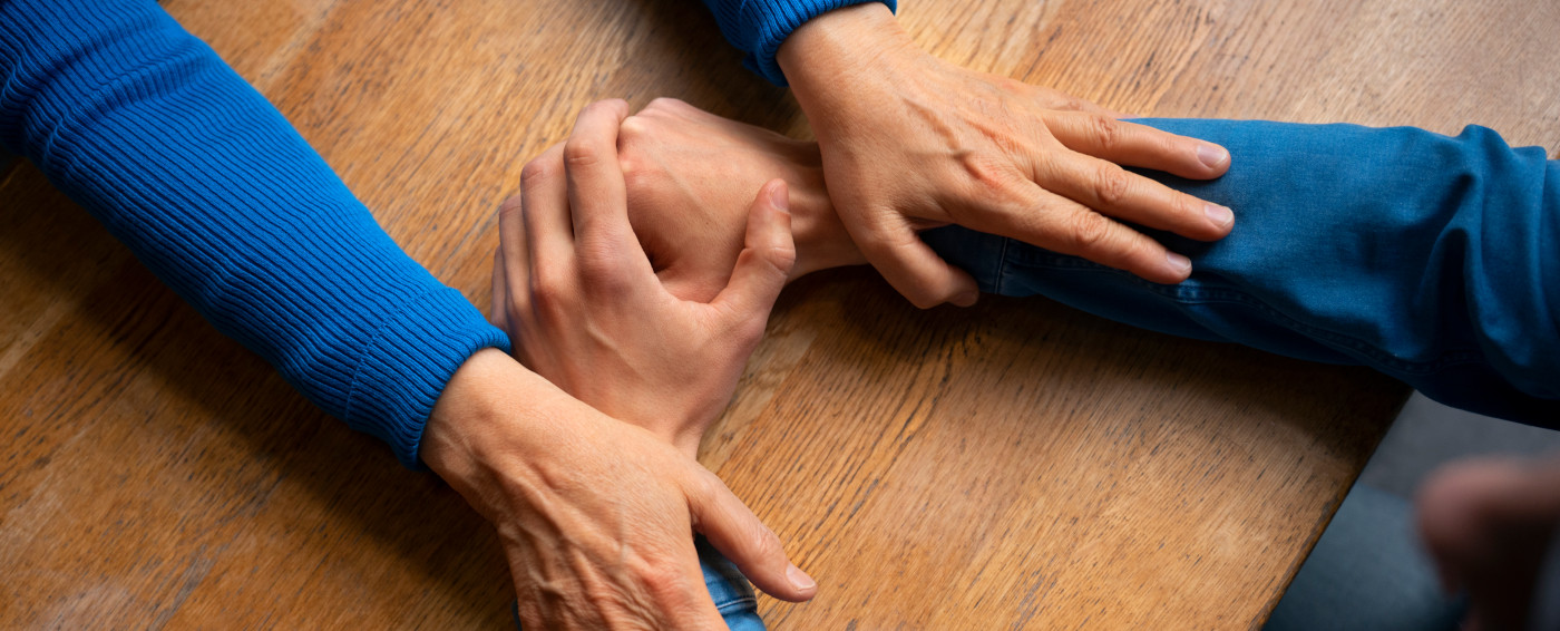 Aparecen las manos de una persona entrelazadas reposando encima de una mesa de madera y, a su vez, las manos de otra persona apoyadas encima de los antebrazos
